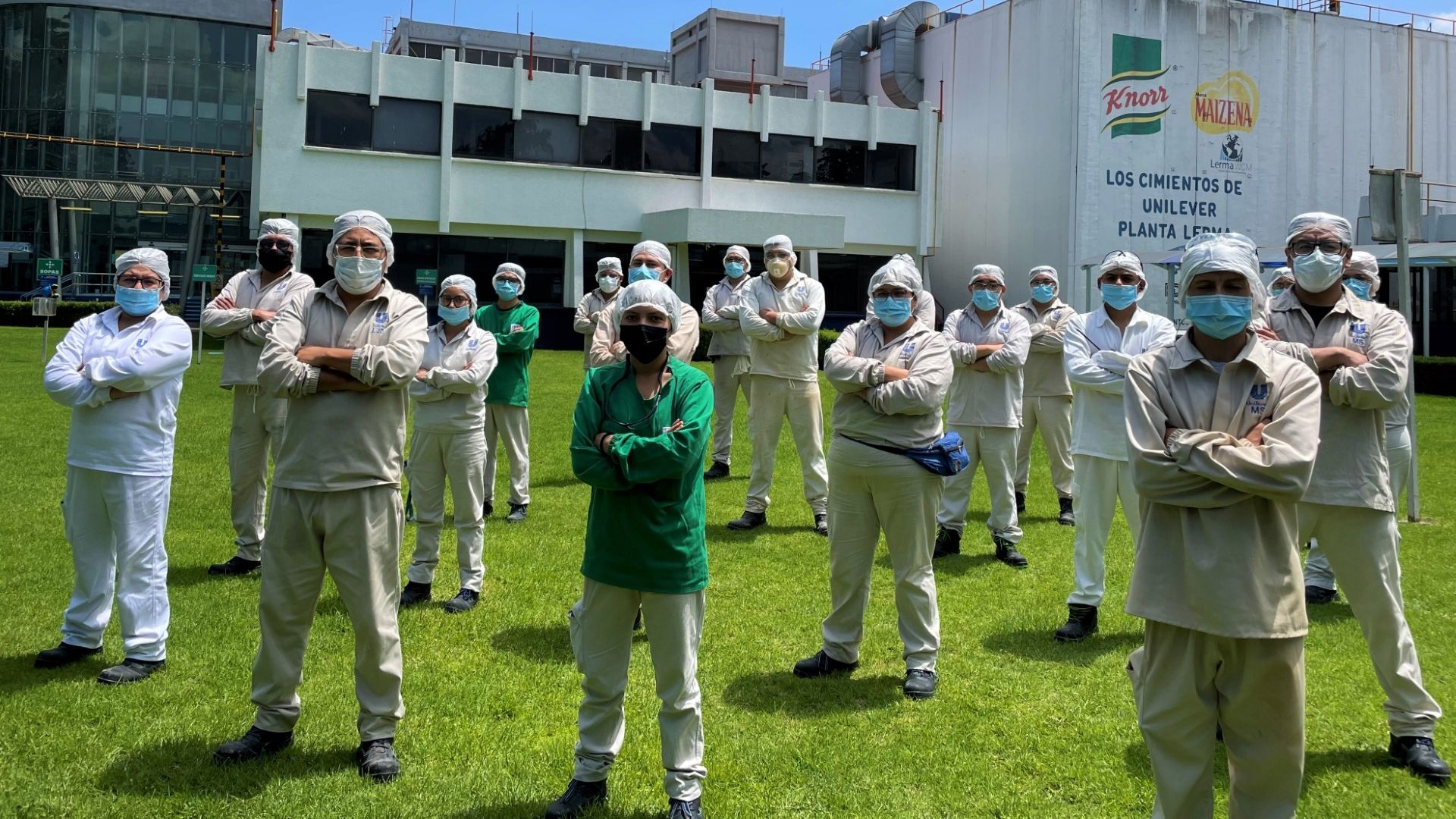 Empleados de una fábrica de Unilever con ropa protectora y mascarillas posan juntos sobre el césped.